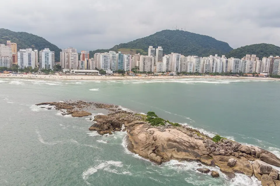 Praia das Astúrias, no Guarujá, com costão rochoso, mar verde e prédios da orla