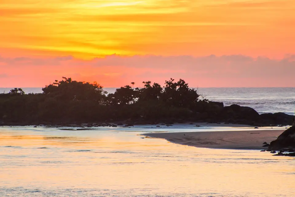 Pôr do sol na Praia da Saudade, em Itanhaém, com rochas e vegetação