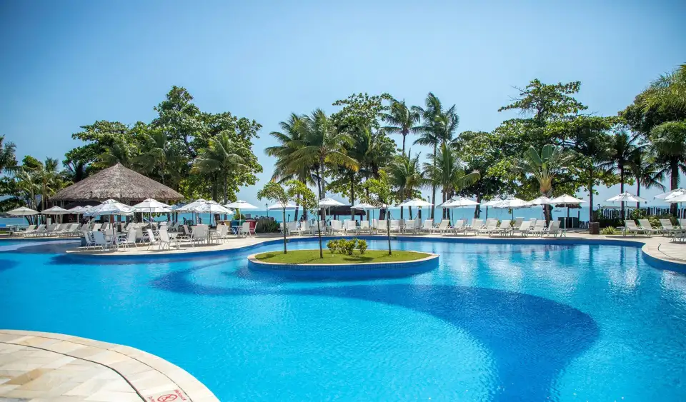 Piscina com ilha de palmeiras do Hotel Jequitimar, com guarda-sóis e o mar ao fundo